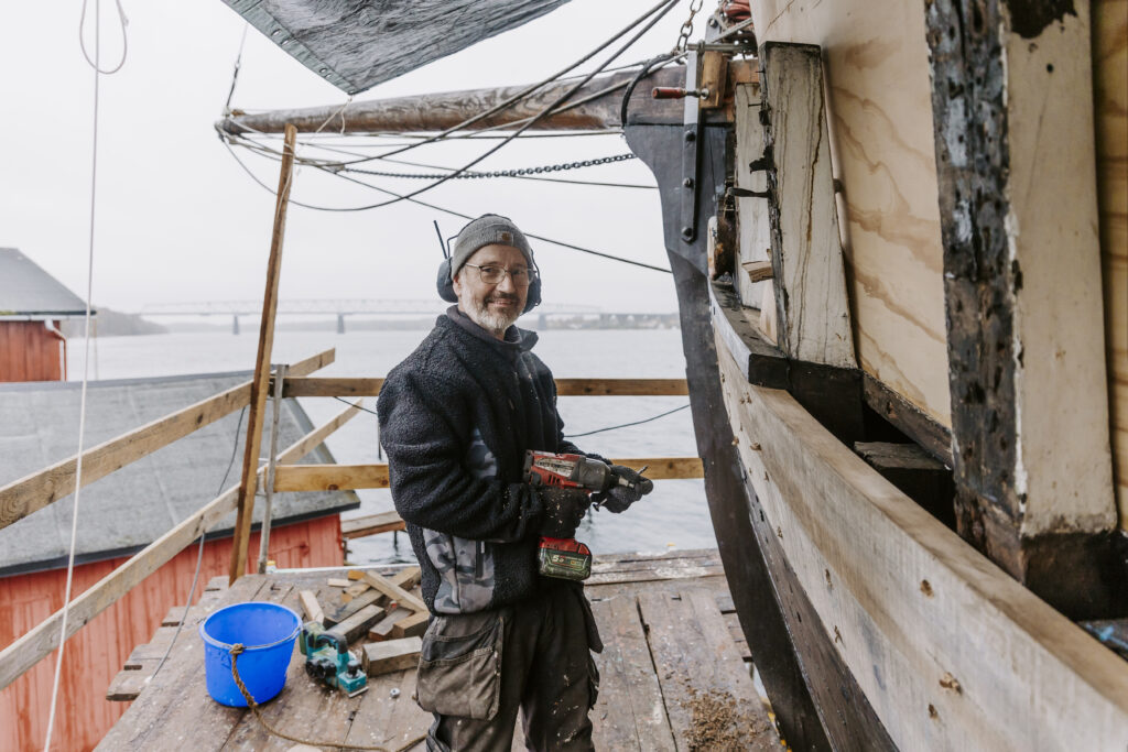 En bådebygger er igang med at renovere et stort træskib. Lillebælt ses i baggrunden.