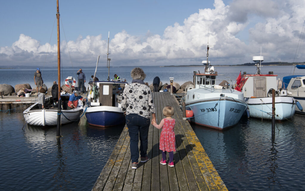 Fiskerjoller ved fiskerleje i Føns med udsigt over hav. En kvinde og en pige går på broen.